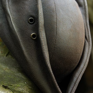 May include: A close-up of a brown leather hat with a textured surface. The hat features two metal eyelets and a stitched seam down the center. The hat's brim is folded over, revealing the interior.