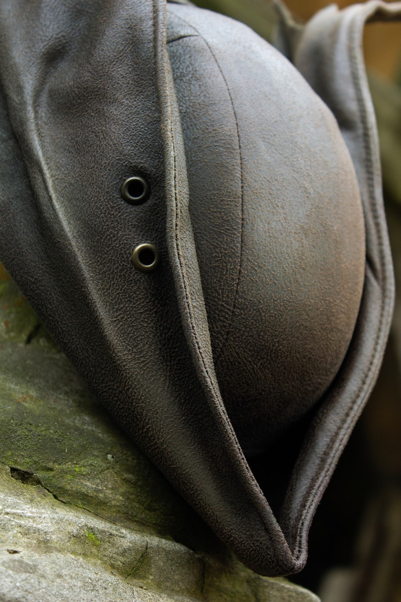 May include: A close-up of a brown leather hat with a textured surface. The hat features two metal eyelets and a stitched seam down the center. The hat's brim is folded over, revealing the interior.