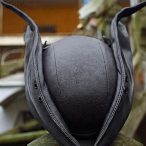 May include: A black leather hat with ear flaps and a rounded crown. The hat has a textured surface and is designed with a stitched seam down the center. The ear flaps extend upwards, giving the hat a unique shape.