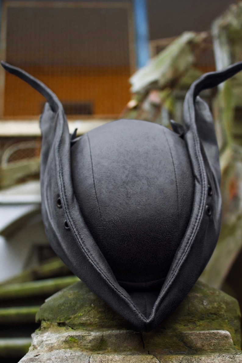 May include: A black leather hat with ear flaps and a rounded crown. The hat has a textured surface and is designed with a stitched seam down the center. The ear flaps extend upwards, giving the hat a unique shape.