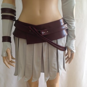 May include: A light gray pleated skirt with a wide, dark burgundy faux leather belt featuring silver-tone metal studs. The set includes matching light gray arm warmers with dark burgundy faux leather straps and silver-tone metal buckles.