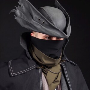 May include: A dark gray, leather-like hat with two eyelets and a torn brim, paired with a black face covering and a black coat with white stitching. A green scarf and chain complete the ensemble.