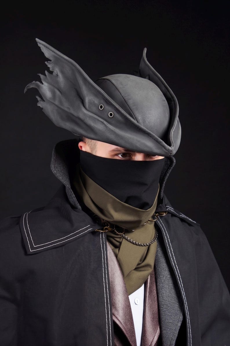May include: A dark gray, leather-like hat with two eyelets and a torn brim, paired with a black face covering and a black coat with white stitching. A green scarf and chain complete the ensemble.