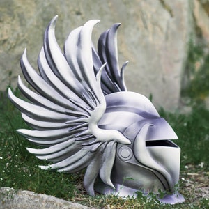May include: A silver helmet with a winged design. The helmet is made of a hard material and has a visor. The helmet is lying on the ground with grass and rocks in the background.