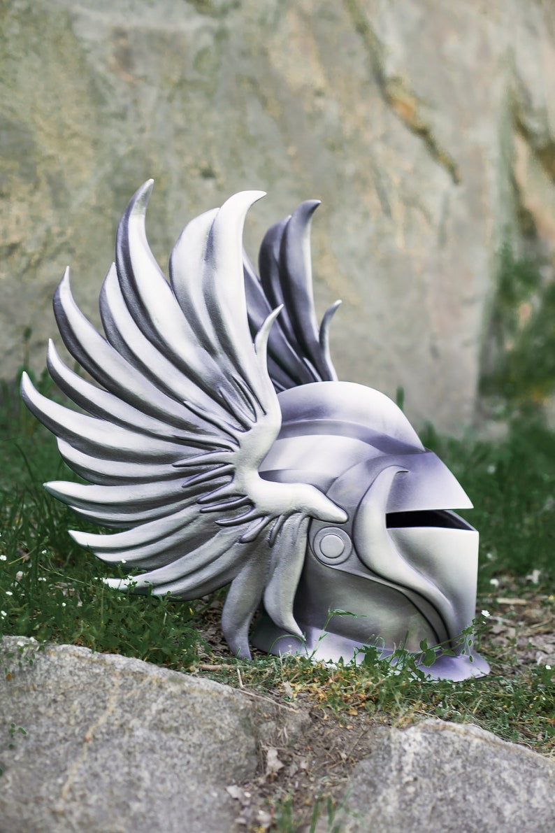 May include: A silver helmet with a winged design. The helmet is made of a hard material and has a visor. The helmet is lying on the ground with grass and rocks in the background.