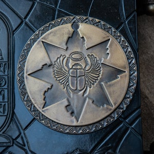 Puede incluir: Primer plano de un diario de cuero negro con un emblema circular de lat&oacute;n que presenta un escarabajo con alas y un halo. El emblema est&aacute; rodeado por una estrella de seis puntas y un borde decorativo.
