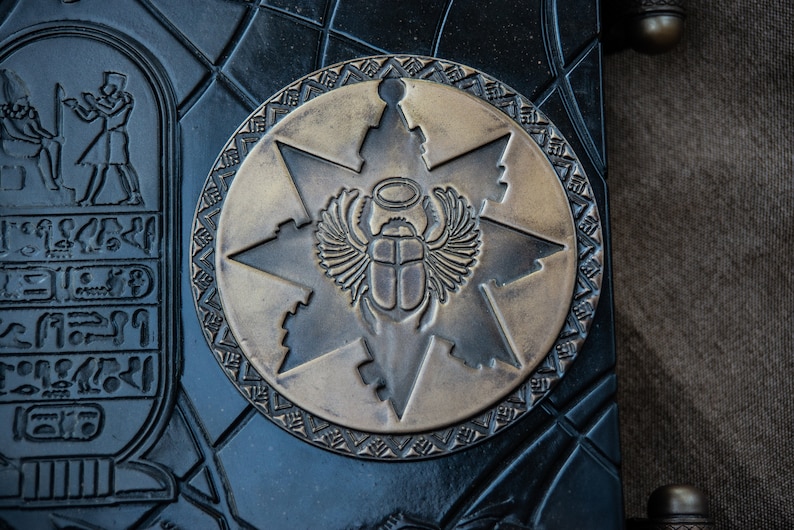 Puede incluir: Primer plano de un diario de cuero negro con un emblema circular de lat&oacute;n que presenta un escarabajo con alas y un halo. El emblema est&aacute; rodeado por una estrella de seis puntas y un borde decorativo.