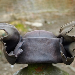 May include: A dark brown leather hat with ear flaps. The hat has a rounded crown and the ear flaps are folded up. The hat is sitting on a green and brown post. The background is blurred.