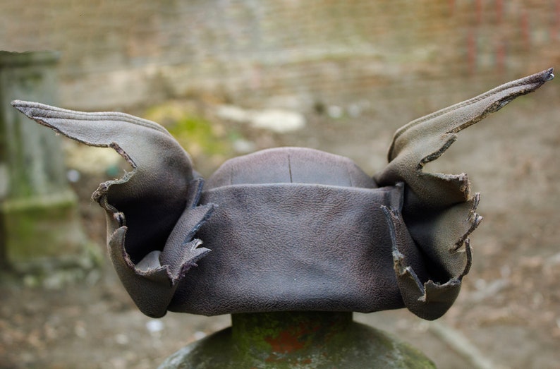 May include: A dark brown leather hat with ear flaps. The hat has a rounded crown and the ear flaps are folded up. The hat is sitting on a green and brown post. The background is blurred.