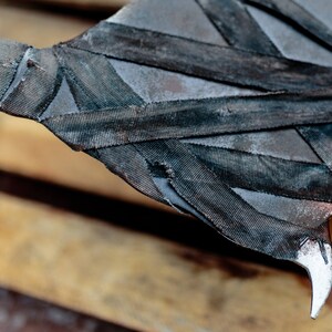 Op de afbeelding: Een metalen bijlkop met een donkere, verweerde afwerking en scherpe, puntige randen. Het oppervlak is gedeeltelijk omwikkeld met stroken zwarte, getextureerde stof die over het metaal kruisen, wat zorgt voor een gelaagd en robuust uiterlijk.
