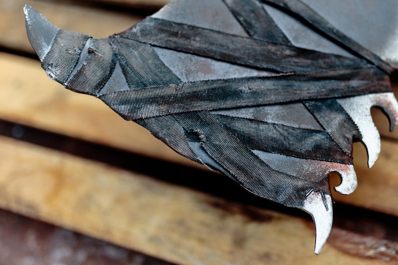 Op de afbeelding: Een metalen bijlkop met een donkere, verweerde afwerking en scherpe, puntige randen. Het oppervlak is gedeeltelijk omwikkeld met stroken zwarte, getextureerde stof die over het metaal kruisen, wat zorgt voor een gelaagd en robuust uiterlijk.