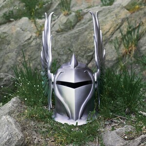 May include: A silver knight helmet with winged accents, resting on a bed of green grass and rocks.