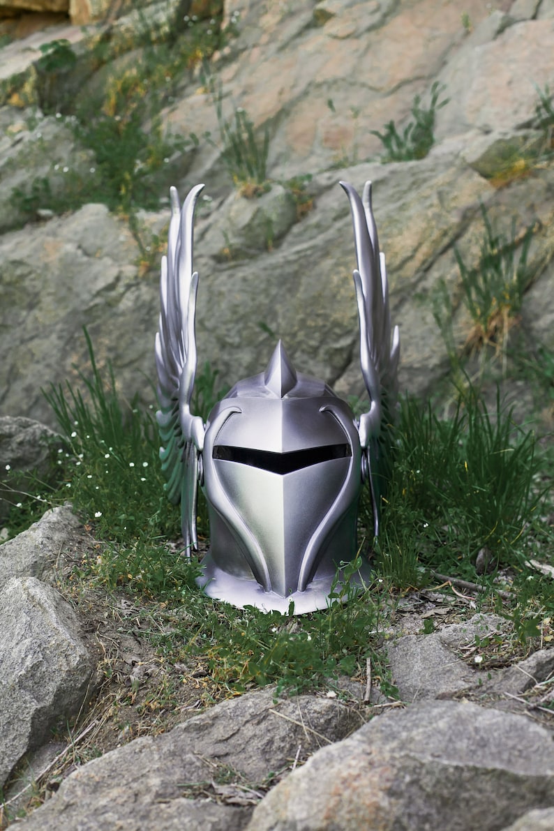 May include: A silver knight helmet with winged accents, resting on a bed of green grass and rocks.