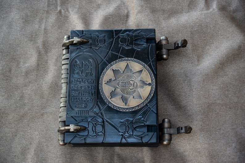 Puede incluir: Un diario encuadernado en cuero negro con intrincados tallados de escarabajos e  jerogl&iacute;ficos. La portada presenta un emblema circular de lat&oacute;n con un dise&ntilde;o de estrella y un escarabajo en el centro.