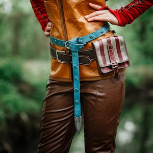 May include: A brown leather belt with a blue and silver buckle and a striped fabric pouch attached. The belt is worn over a brown leather jacket.