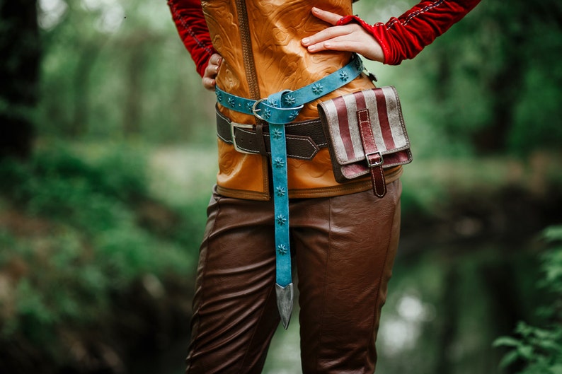 May include: A brown leather belt with a blue and silver buckle and a striped fabric pouch attached. The belt is worn over a brown leather jacket.