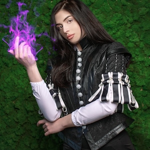 May include: A woman in a black leather jacket with white and black trim, holding a purple flame in her hand. She is standing in front of a green moss wall.