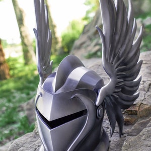 May include: A silver helmet with a winged design. The helmet has a pointed top and a face shield. The wings are detailed and extend out from the sides of the helmet.
