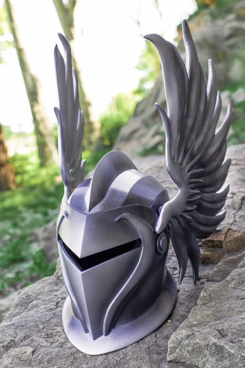 May include: A silver helmet with a winged design. The helmet has a pointed top and a face shield. The wings are detailed and extend out from the sides of the helmet.