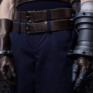 May include: A close-up of a person wearing a dark blue outfit with a brown leather belt and two large, metal gauntlets on their arms.