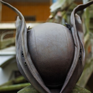 May include: A brown leather hat with a unique design, featuring a rounded crown and wide, upturned brims. The hat has visible stitching and eyelets, suggesting a vintage or historical style. The hat is resting on a stone surface.