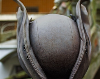 May include: A brown leather hat with a unique design, featuring a rounded crown and wide, upturned brims. The hat has visible stitching and eyelets, suggesting a vintage or historical style. The hat is resting on a stone surface.