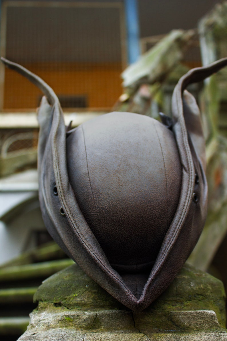 May include: A brown leather hat with a unique design, featuring a rounded crown and wide, upturned brims. The hat has visible stitching and eyelets, suggesting a vintage or historical style. The hat is resting on a stone surface.