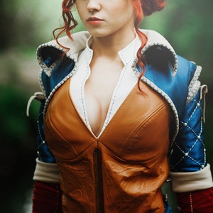 May include: A woman wearing a brown leather vest with a blue and white patterned jacket over it. The vest has a deep V-neckline and is fastened with a zipper.