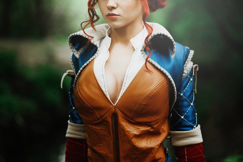 May include: A woman wearing a brown leather vest with a blue and white patterned jacket over it. The vest has a deep V-neckline and is fastened with a zipper.