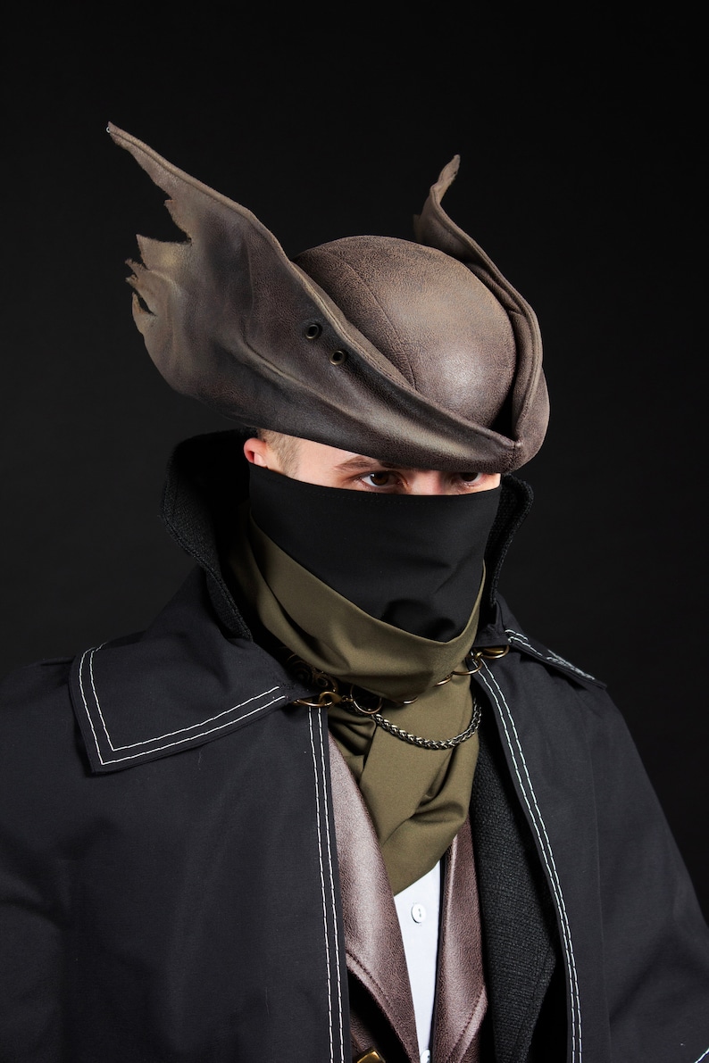 May include: A cosplay outfit featuring a brown leather hat with torn edges, a black face covering, and a black coat with white stitching. The outfit includes a green scarf and a chain necklace.