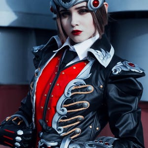 Puede incluir: Un disfraz de cosplay con una chaqueta negra con detalles de cuerda dorada y un chaleco rojo. El atuendo incluye un tocado plateado y rojo y accesorios para los brazos a juego. La persona lleva maquillaje oscuro y lápiz labial rojo.