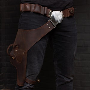 May include: Brown leather holster and belt set. The holster is designed to be worn on the thigh and is attached to a belt with a large silver buckle. The belt has multiple loops for ammunition. The leather has a distressed appearance.