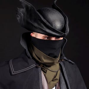 May include: A black, leather-like hat with pointed, wing-like accents. The hat is part of a costume ensemble, including a black face covering, a dark green scarf, and a black coat with white stitching. A chain necklace adds detail to the outfit.
