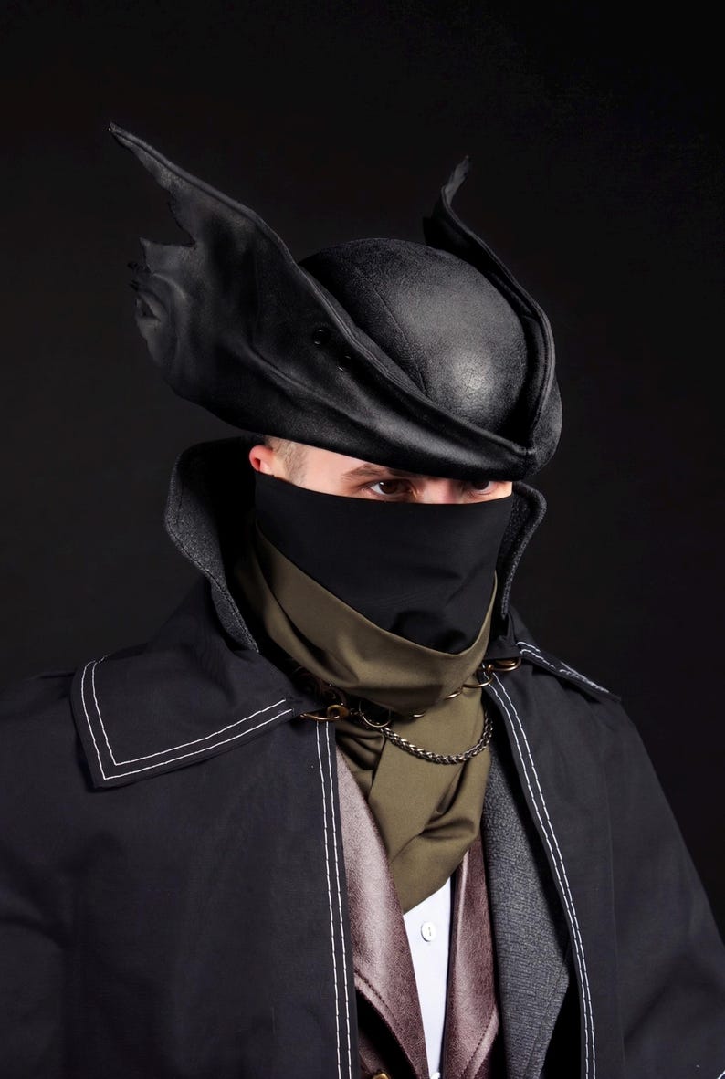 May include: A black, leather-like hat with pointed, wing-like accents. The hat is part of a costume ensemble, including a black face covering, a dark green scarf, and a black coat with white stitching. A chain necklace adds detail to the outfit.