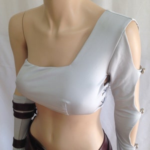 May include: A light gray one-shoulder crop top featuring a long sleeve with cutouts secured by silver-toned metal buttons. The garment includes side lacing details and is paired with a dark maroon belt and a matching gray forearm sleeve with straps.