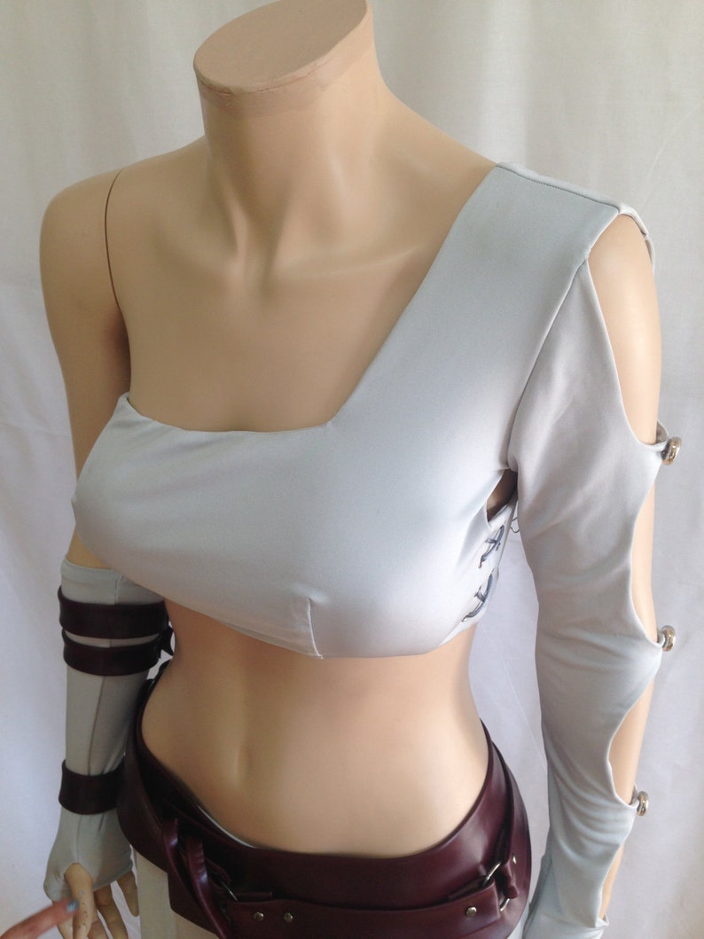 May include: A light gray one-shoulder crop top featuring a long sleeve with cutouts secured by silver-toned metal buttons. The garment includes side lacing details and is paired with a dark maroon belt and a matching gray forearm sleeve with straps.