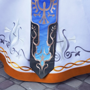 May include: A white skirt with a gold and orange trim. The skirt features a blue and gold embroidered design with a stylised bird.