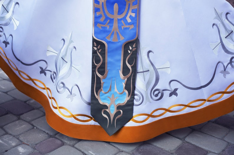 May include: A white skirt with a gold and orange trim. The skirt features a blue and gold embroidered design with a stylised bird.