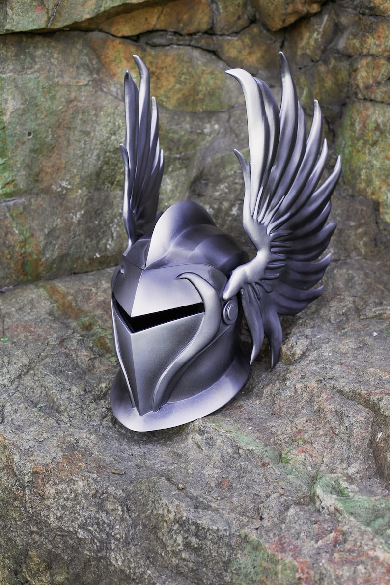 May include: A silver helmet with a winged design. The helmet has a pointed visor and a textured surface. The wings are detailed and have a feathered appearance.