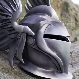 May include: A silver helmet with a winged design. The helmet has a visor and a textured surface.
