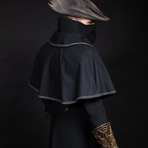 May include: A dark, gothic-style costume featuring a black cape with white stitching, a high-collared shirt, and a unique, weathered hat. The outfit includes a decorative gold arm guard and black gloves, set against a dark background.