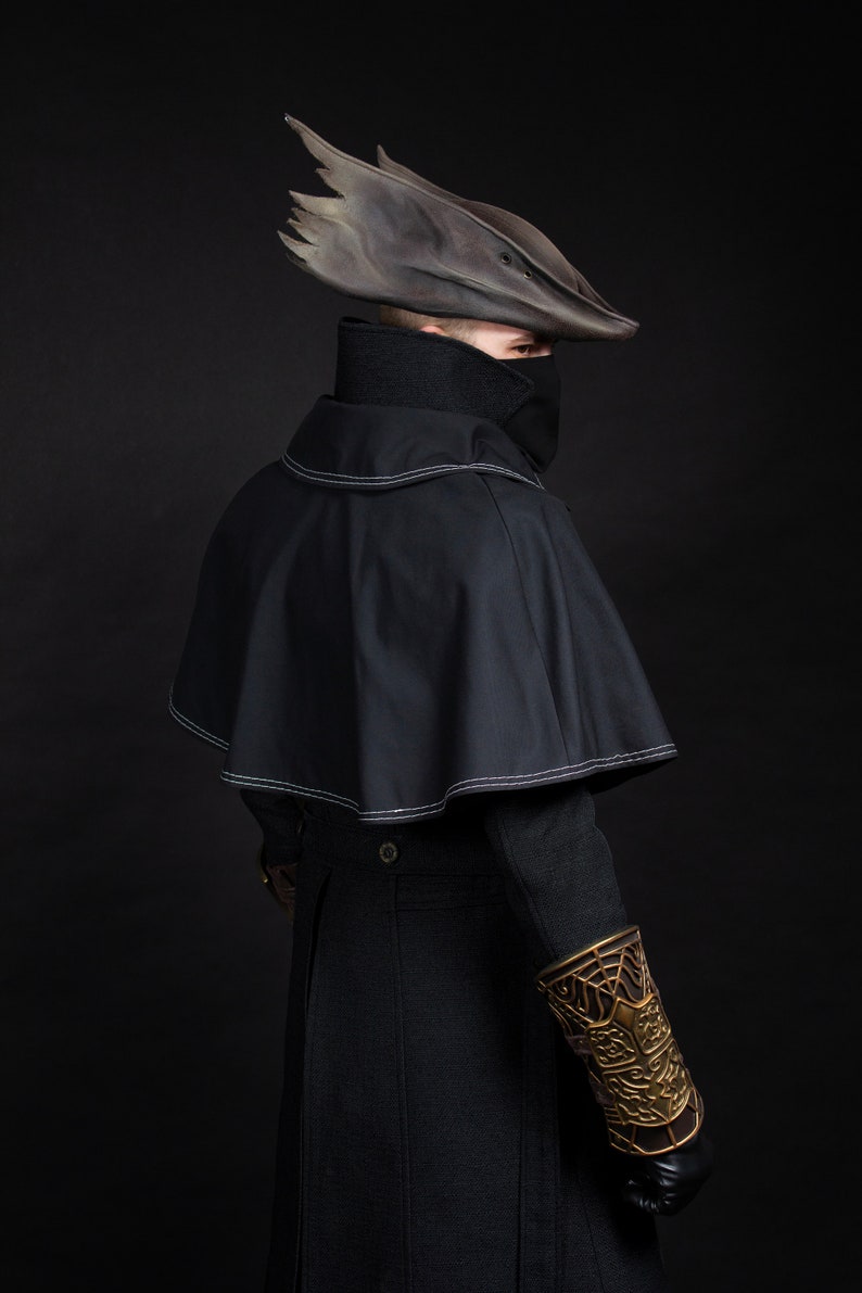 May include: A dark, gothic-style costume featuring a black cape with white stitching, a high-collared shirt, and a unique, weathered hat. The outfit includes a decorative gold arm guard and black gloves, set against a dark background.