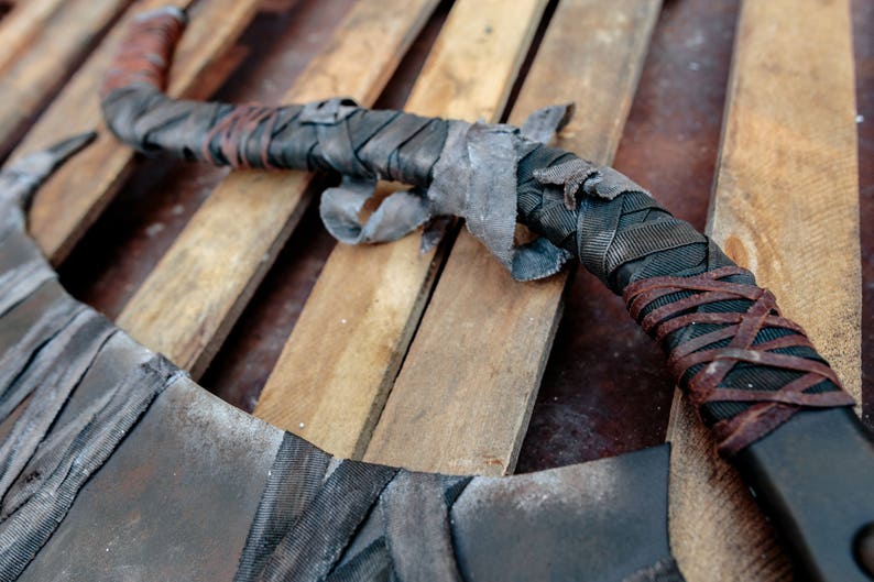 Op de afbeelding: Een gebogen wapen met een lang handvat dat is omwikkeld met lagen zwarte stoffen tape en dunne bruine leren stroken. Het metalen lemmet heeft een verweerde, donkergrijze afwerking met zichtbare tekenen van slijtage op het oppervlak.
