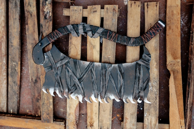 Op de afbeelding: Een groot, gebogen hakmes met een donkere, verweerde metalen afwerking en een gekartelde onderkant. Het handvat en het blad zijn omwikkeld met stroken donkere, versleten stof, vastgezet met dunne bruine koordaccenten voor een textuur.