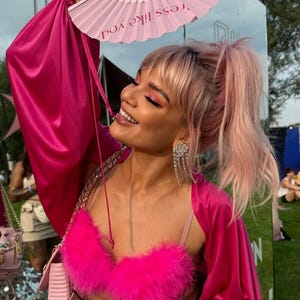 May include: A woman wearing a bright pink outfit, including a feathered bra, satin jacket, and a pink fan with the text "dress like you". She has pink hair and is wearing dangling rhinestone earrings.