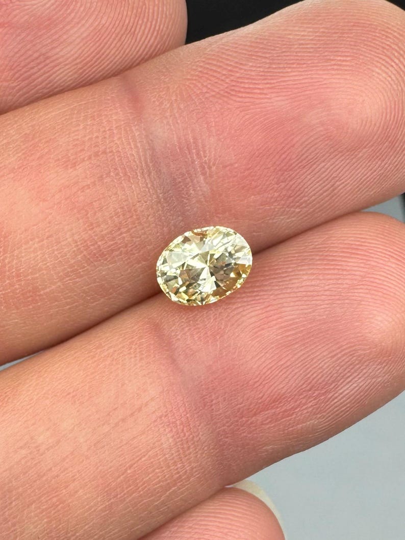 以下が含まれることがあります： 明るい黄色のオーバルカットダイヤモンド。宝石は指の間に挟まれ、その輝きと透明度を示しています。ダイヤモンドのファセットは光を反射し、きらめく効果を生み出します。全体的な色は淡い金色です。