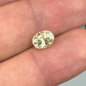 以下が含まれることがあります： 薄い黄色の楕円形にカットされたダイヤモンド。宝石は指の間に挟まれ、そのファセットと透明度を強調しています。ダイヤモンドのカットは光を反射し、きらめく効果を生み出します。全体的なテーマはジュエリーと宝石です。