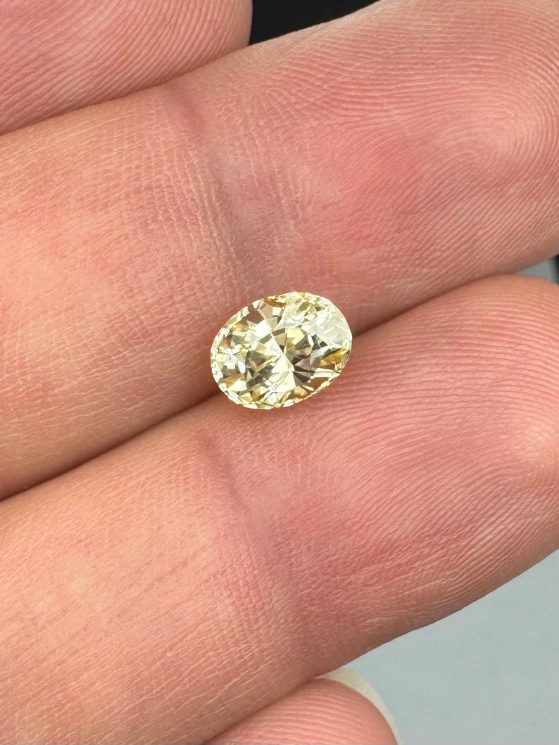 以下が含まれることがあります： 薄い黄色の楕円形にカットされたダイヤモンド。宝石は指の間に挟まれ、そのファセットと透明度を強調しています。ダイヤモンドのカットは光を反射し、きらめく効果を生み出します。全体的なテーマはジュエリーと宝石です。
