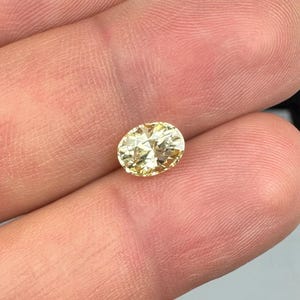 以下が含まれることがあります： 楕円形、淡い黄色の宝石。 宝石は指の間に挟まれ、そのファセットと輝きを際立たせています。 石の色は柔らかく淡い黄色で、よく磨かれており、光を反射しているようです。 宝石がメインです。