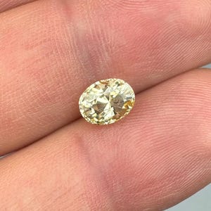 以下が含まれることがあります： 薄い黄色の楕円形にカットされた宝石。 宝石は指の間に挟まれ、その形と色を際立たせています。 石のカットは、きらめく効果を生み出します。 この宝石はジュエリーに使用できます。